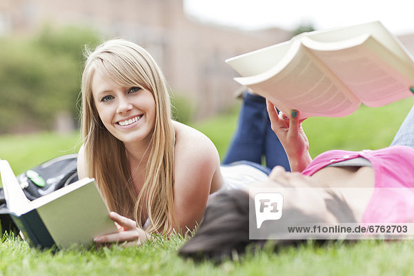 Two female college student lying on grass reading books  one looking at camera