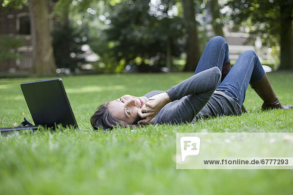 Young woman lying on grass using laptop and cell phone