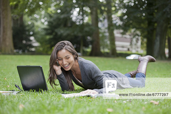 Young woman lying on grass using laptop and cell phone