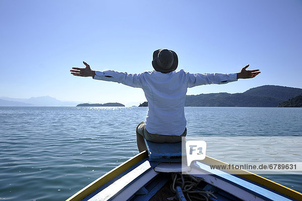Young man sitting on the bow of a fishing boat with his arms wide open  Bay of Paraty or Parati  State of Rio de Janeiro  Brazil  South America