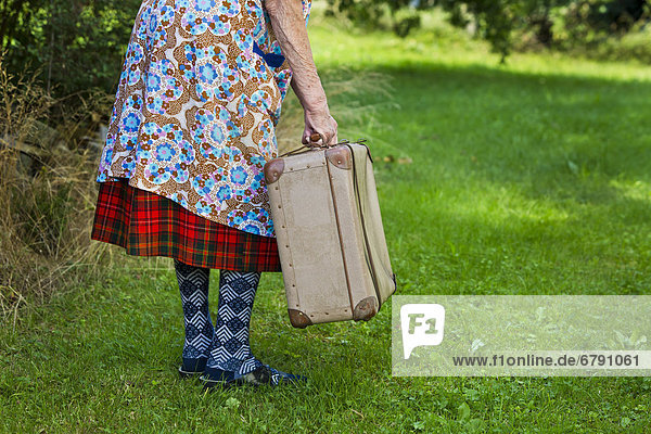 Alte Frau steht mit Koffer in der Wiese