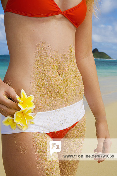 Hawaii  Oahu  Lanikai  Bikini bekleideten Mädchen am Strand hält Plumeria Blumen  Nahaufnahme auf Zwerchfell in Sand bedeckt.