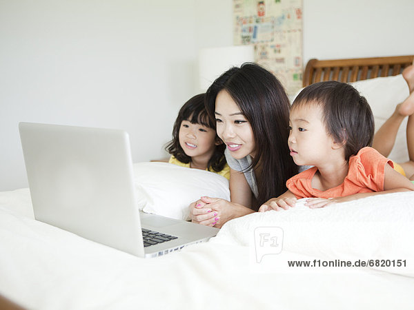 Mother with daughters (2-3  4-5) lying down in bed  using laptop