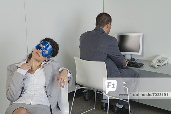 Woman napping with eye mask in office as colleague works nearby