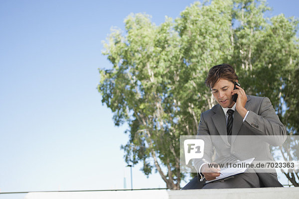Businessman talking on cell phone and writing on clipboard outdoors