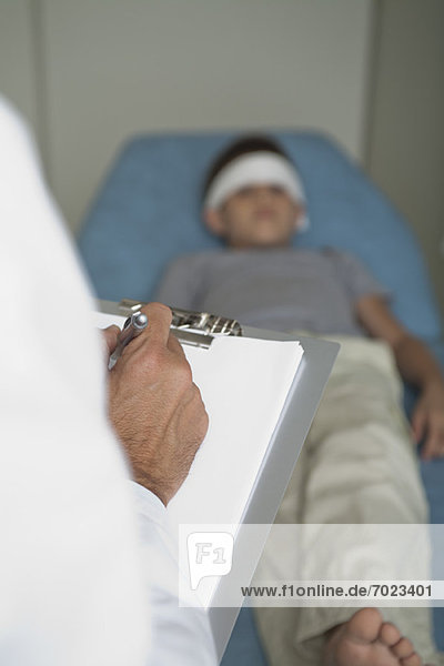 Arzt schreibt auf Klemmbrett  Junge liegt auf Untersuchungstisch mit Stirnbandage im Hintergrund
