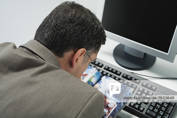 Mature man reading comic book at desk in office  rear view