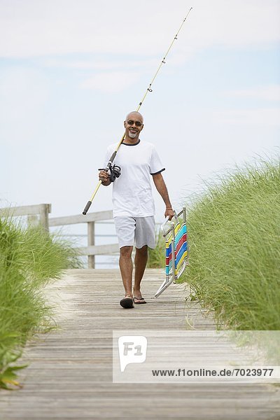 Man with fishing rod and folding chair walking on boardwalk
