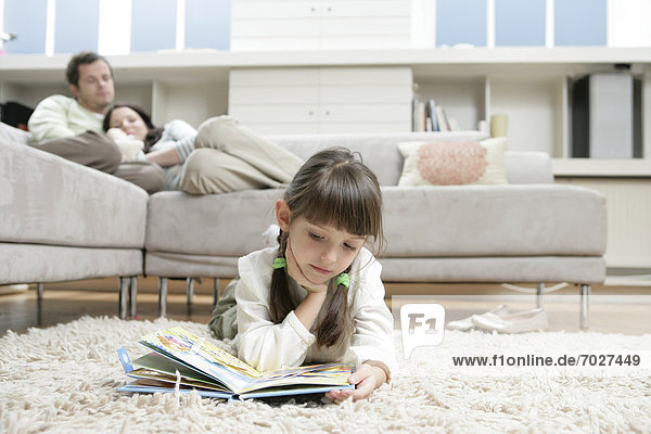 Girl reading book on floor  parents on sofa in background