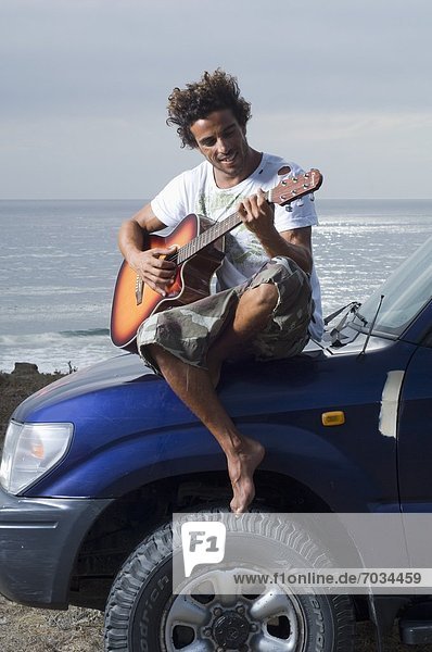 Mid Adult Male Playing Guitar While Sitting On Hood Of Car
