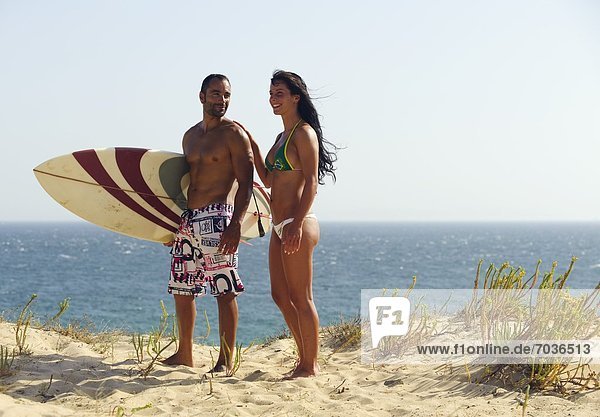 Couple Standing Together On The Beach  With A Surfboard