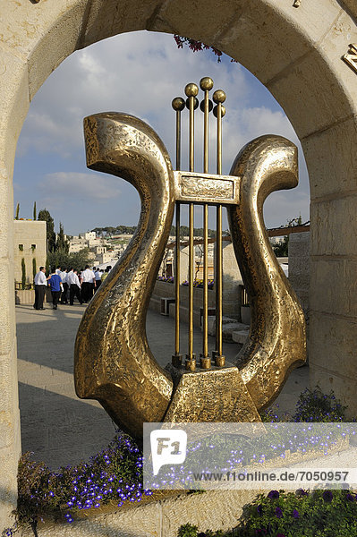 Skulptur einer Davidsharfe am Eingang der Davidstadt einer archäologischen Ausgrabungsstätte dahinter eine jüdische Besuchergruppe und ein palästinensisches Dorf Ostjerusalem Jerusalem Israel Vorderasien Naher Osten