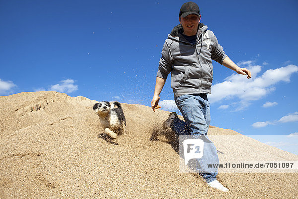 Ein junger Mann mit Basecap rennt zusammen mit einem kleinen Hund (Canis lupus familiaris) einen Sandhügel hinunter  Öttingen  Bayern  Deutschland  Europa
