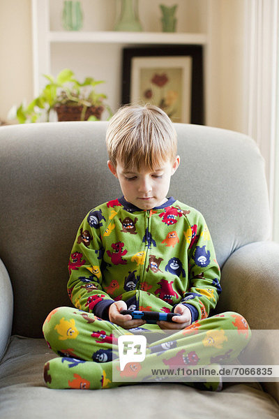 Boy in his pijamas sitting on armchair and playing video game.
