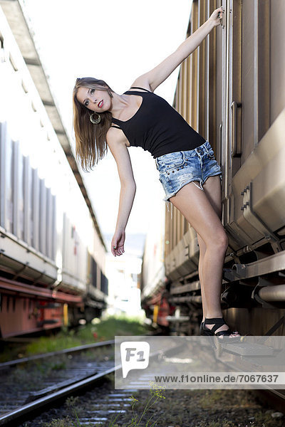 Junge Frau mit schwarzem Top  Jeans-Hotpants und hohen Schuhen posiert zwischen Güterwagons
