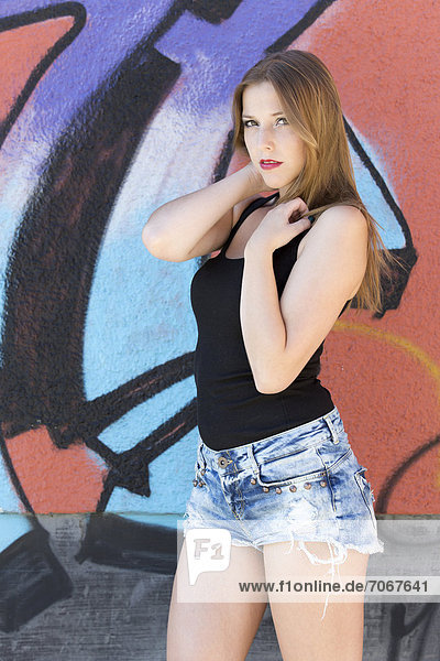 Junge Frau mit schwarzem Top und Jeans-Hotpants posiert vor Wand mit Graffiti
