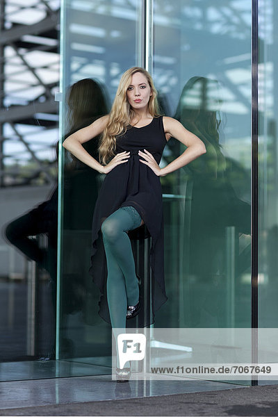 Young woman in a short black dress and green stockings posing in front of a green glass wall