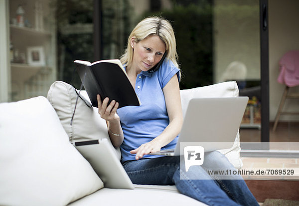 Mittlere erwachsene Frau mit Laptop und Handy  die ein Buch im Wohnzimmer hält.