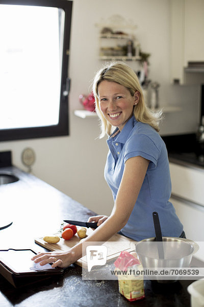 Mittlere erwachsene Frau mit Rezept auf digitalem Tisch und Zubereitung des Essens in der Küche