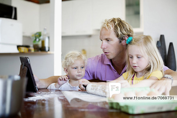 Mann schaut auf digitales Tablett  während kleine Tochter Teig auf dem Tisch in der Küche rollt.