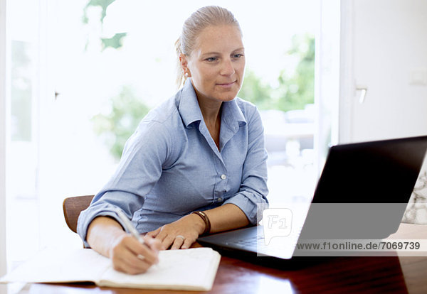 Mid adult woman writing notes while looking at laptop