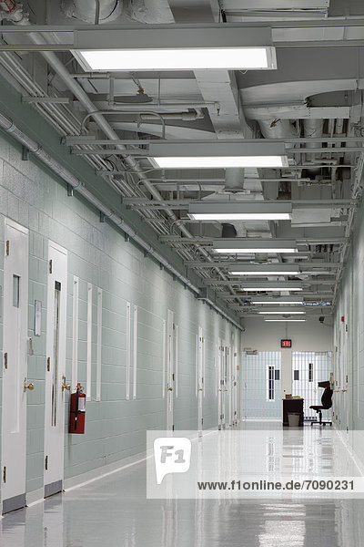 A long corridor in a residential unit at a Correctional Facility. Prison cell doors with observation hatches. Fire extinguisher. Table and chair.