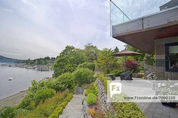 A house terrace overlooking the water  the shore. Decking  plants and outdoor furniture. Boats moored.
