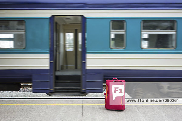 An empty railway station in Estonia. A single red suitcase on the platform beside the open door of a train carriage.