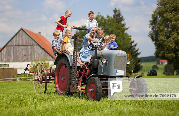 Frau mit Kindergruppe auf altem Traktor vor dem Bauernhaus sitzend