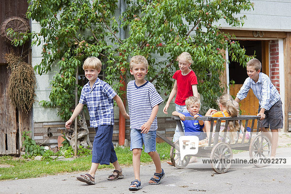 Germany  Bavaria  Group of children playing with hand cart in front of farmhouse
