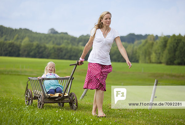 Germany  Bavaria  Mother pulling daughter in hand cart through meadow