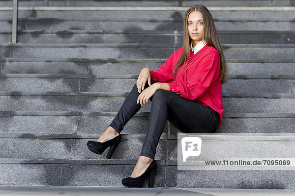 Young woman wearing a red top  black jeans and high heels posing while sitting on stairs