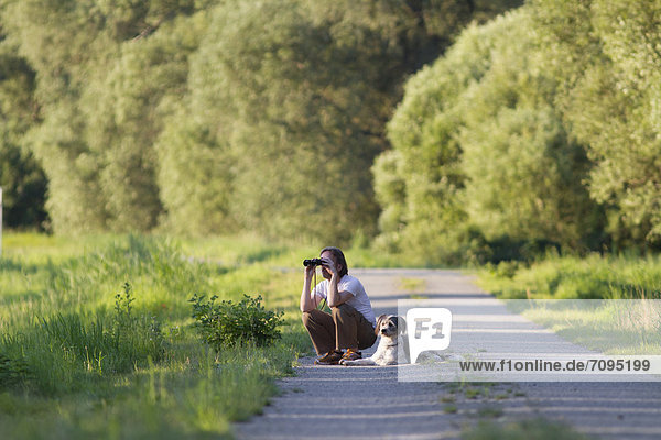 Mann mit Hund hockt auf einem Weg und beobachtet etwas mit dem Fernglas  Wustermark  Brandenburg  Deutschland  Europa