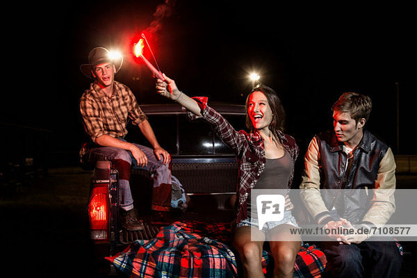 Three friends sitting on tailgate of car at night  girl holding sparkler