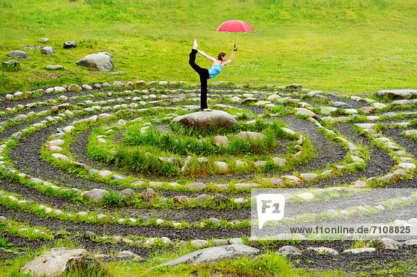 Frau hält einen Regenschirm  während sie im Yoga in der Mitte des Steinlabyrinths posiert.