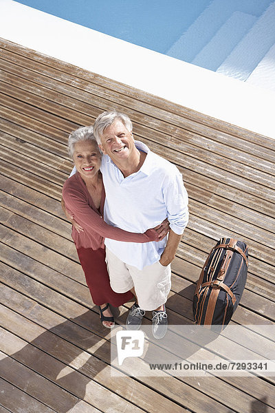 Spain  Senior couple standing with suitcase at swimming pool  smiling  portrait