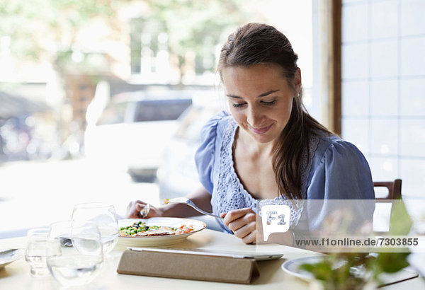 Junge Frau beim Essen  während sie digitale Tabletten am Restauranttisch benutzt.