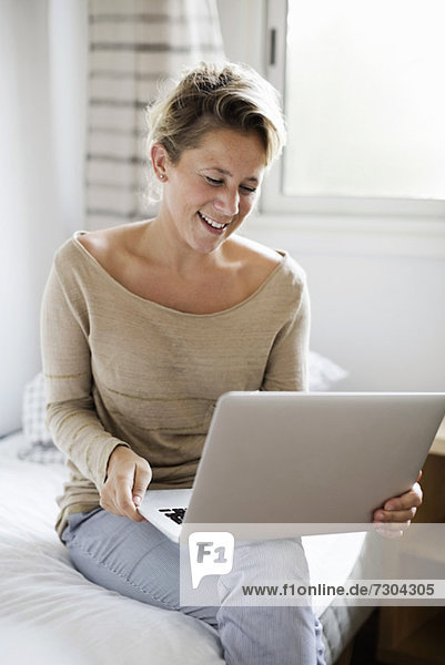 Glückliche mittlere erwachsene Frau mit Laptop im Bett zu Hause