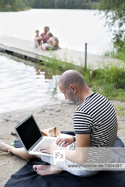 Erwachsener Mann mit Laptop am Strand mit Familie auf dem Pier sitzend