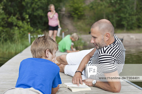 Erwachsener Mann mit Sohn auf dem Pier liegend und Familie im Hintergrund stehend