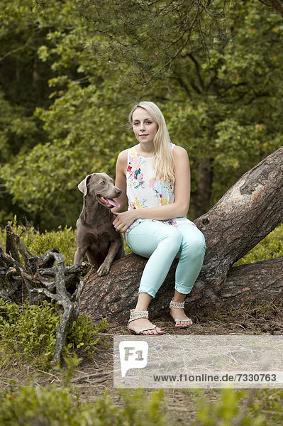 Frau sitzt mit einem Labrador Retriever auf einem Baumstamm
