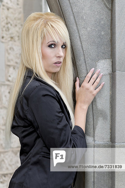Junge Frau mit langen blonden Haaren und schwarzer Jacke posiert an Mauerbogen