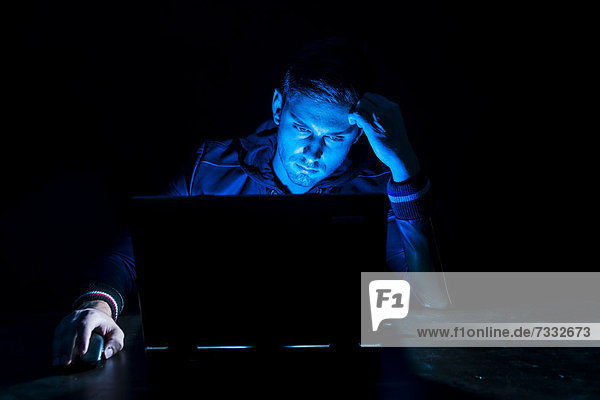 Junger Mann sitzt im Dunkeln vor seinem Laptop  nur vom Monitor angestrahlt