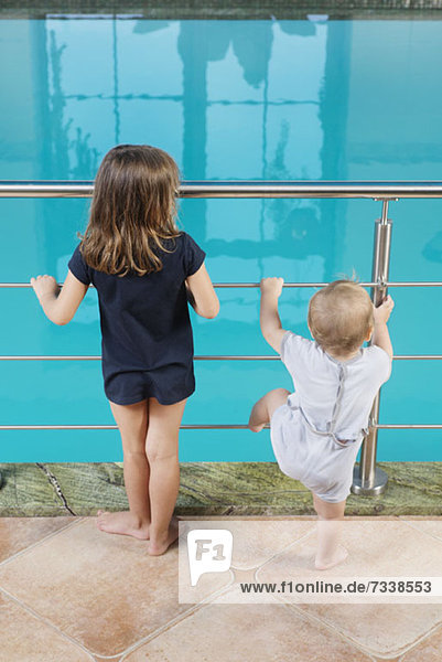 Ein junges Mädchen und ihr kleiner Bruder stehen nebeneinander und schauen in ein Schwimmbad.