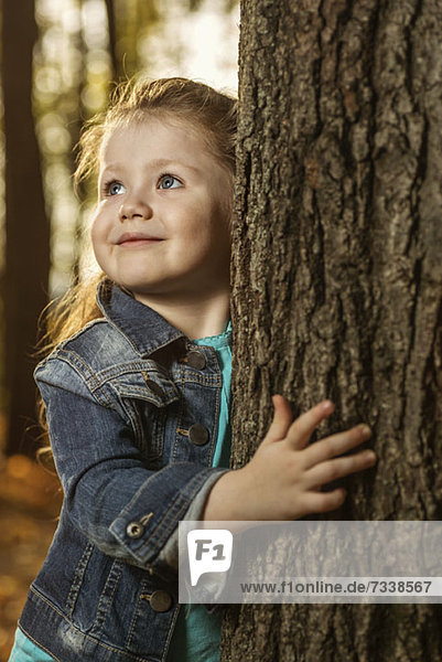 Ein junges Mädchen hält sich an einem Baumstamm fest und schaut neugierig weg.