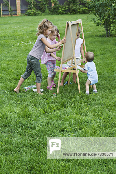 Drei junge Mädchen und ein Kleinkind malen im Freien auf einer Staffelei.