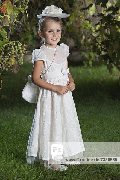 A smiling young girl dressed elegantly in a white dress  hat and purse