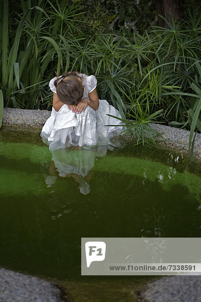 Ein junges trauriges Mädchen  das seinen Kopf auf die Hände legt  während es an einem Teich sitzt.