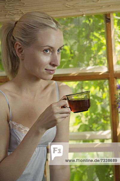 A beautiful woman in a nightie sitting by a window drinking tea