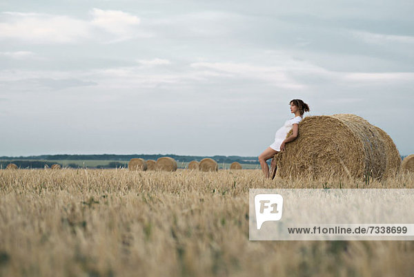 A pregnant woman leaning against a bale of hay in a remote setting